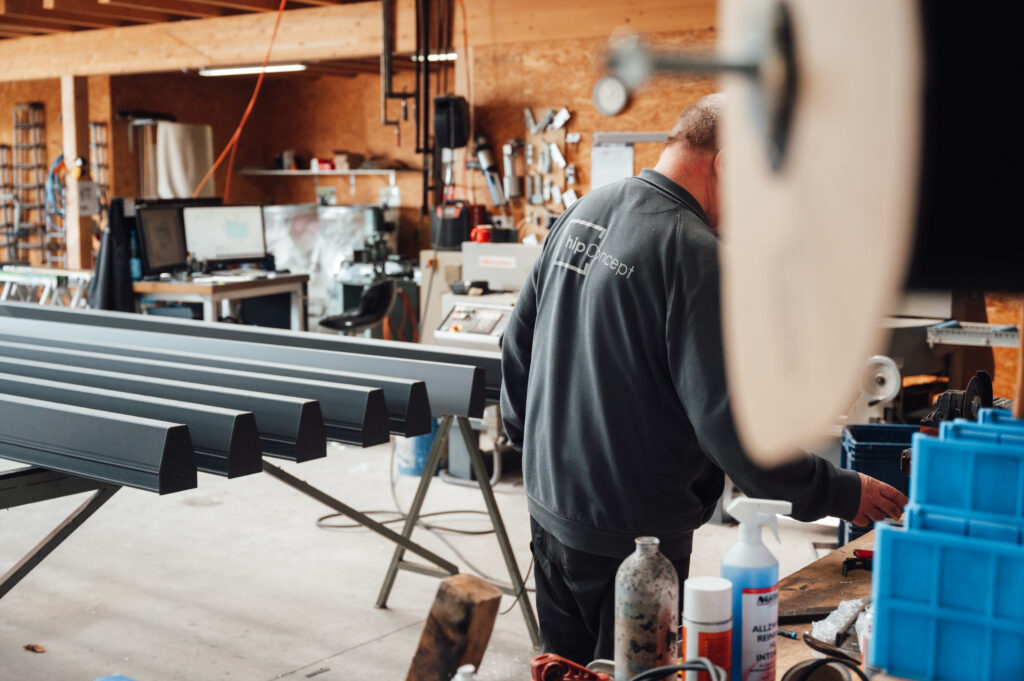 Es wird ein Aufschnitt aus der Fertigungshalle gezeigt. Auf zwei Böcken liegen Aluminiumprofile und man erkennt einen Mitarbeiter aus der Produktion